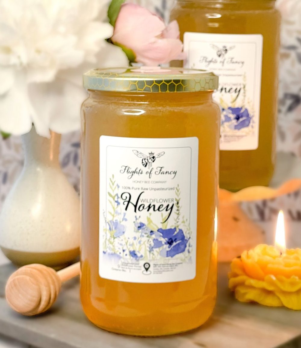 A large jar of their wildflower honey beside a honey dipper and a lit beeswax flower candle.