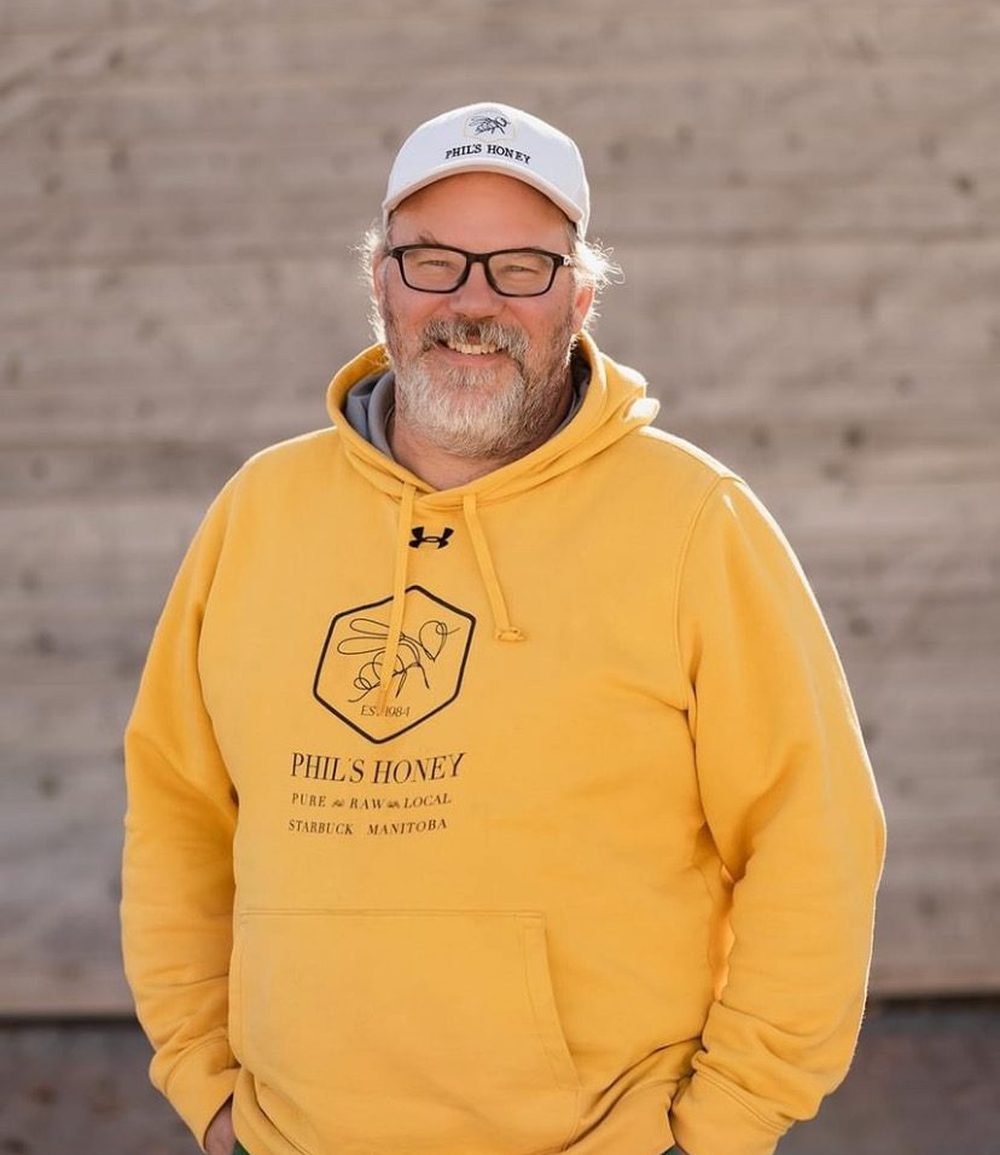 Phil Veldhuis outside a weathered wooden wall, in a yellow Phil's Honey hoodie and cap.